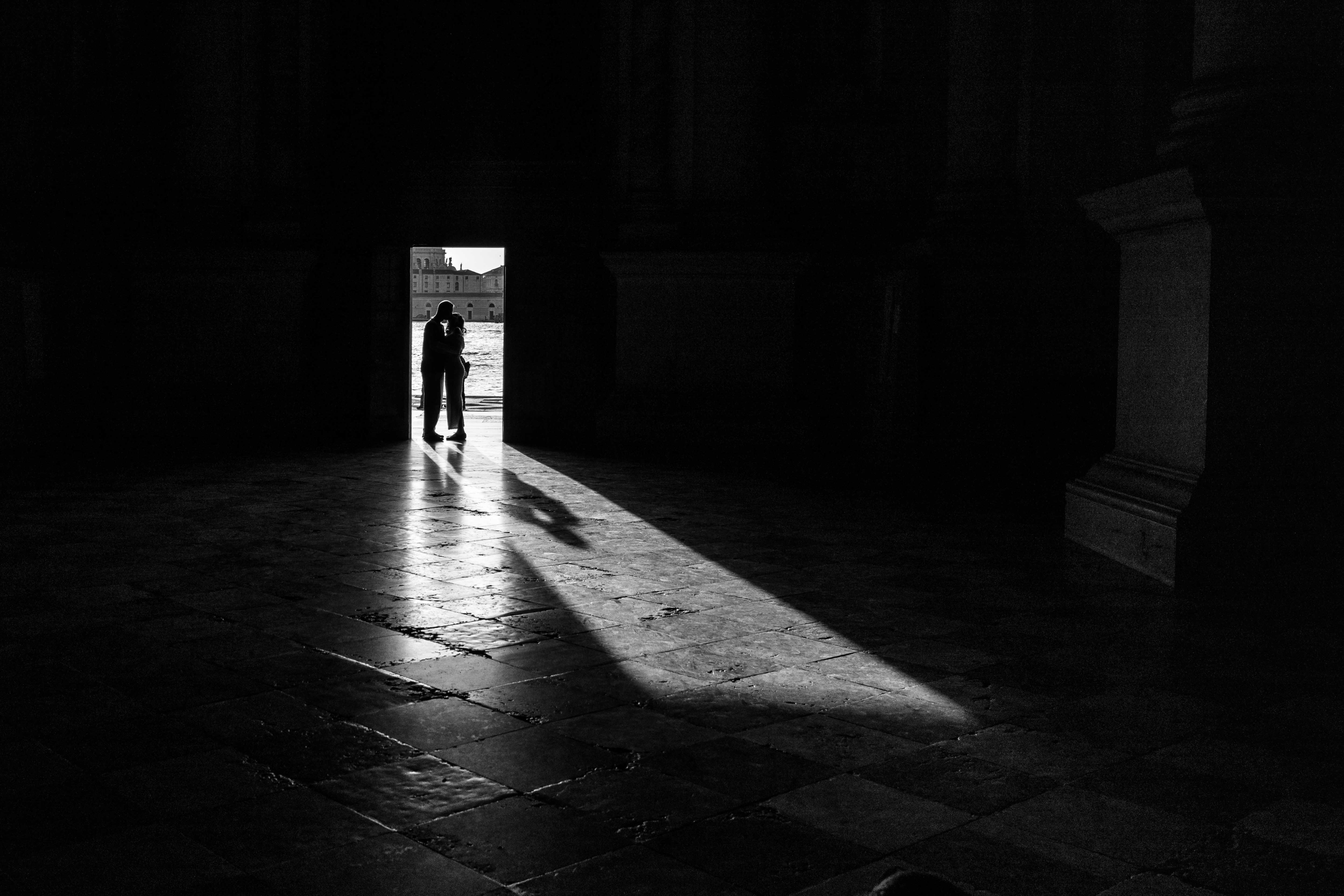 Black-and-white silhouette of a couple embracing in a sunlit doorway at San Giorgio Maggiore, Venice, with a shaft of light stretching across the stone floor